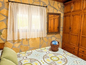 A room with a stone wall, a wooden wardrobe, a bed with patterned bedding, and a window with sheer curtains. - Juan De Dios Farmhouse (Cómpeta)