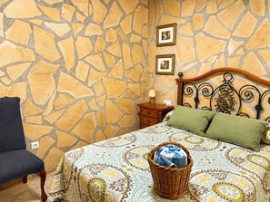 Ein Schlafzimmer mit einer Wand im Steinmuster, einem Bett mit gemusterten Betttextilien, einem Nachttisch, einer Lampe und einem Sessel.