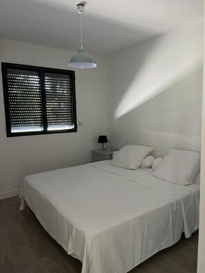 A bedroom with a bed, a nightstand, a lamp, and a window with blinds. - Villa Seignosse: A Beach Vacation (Seignosse)