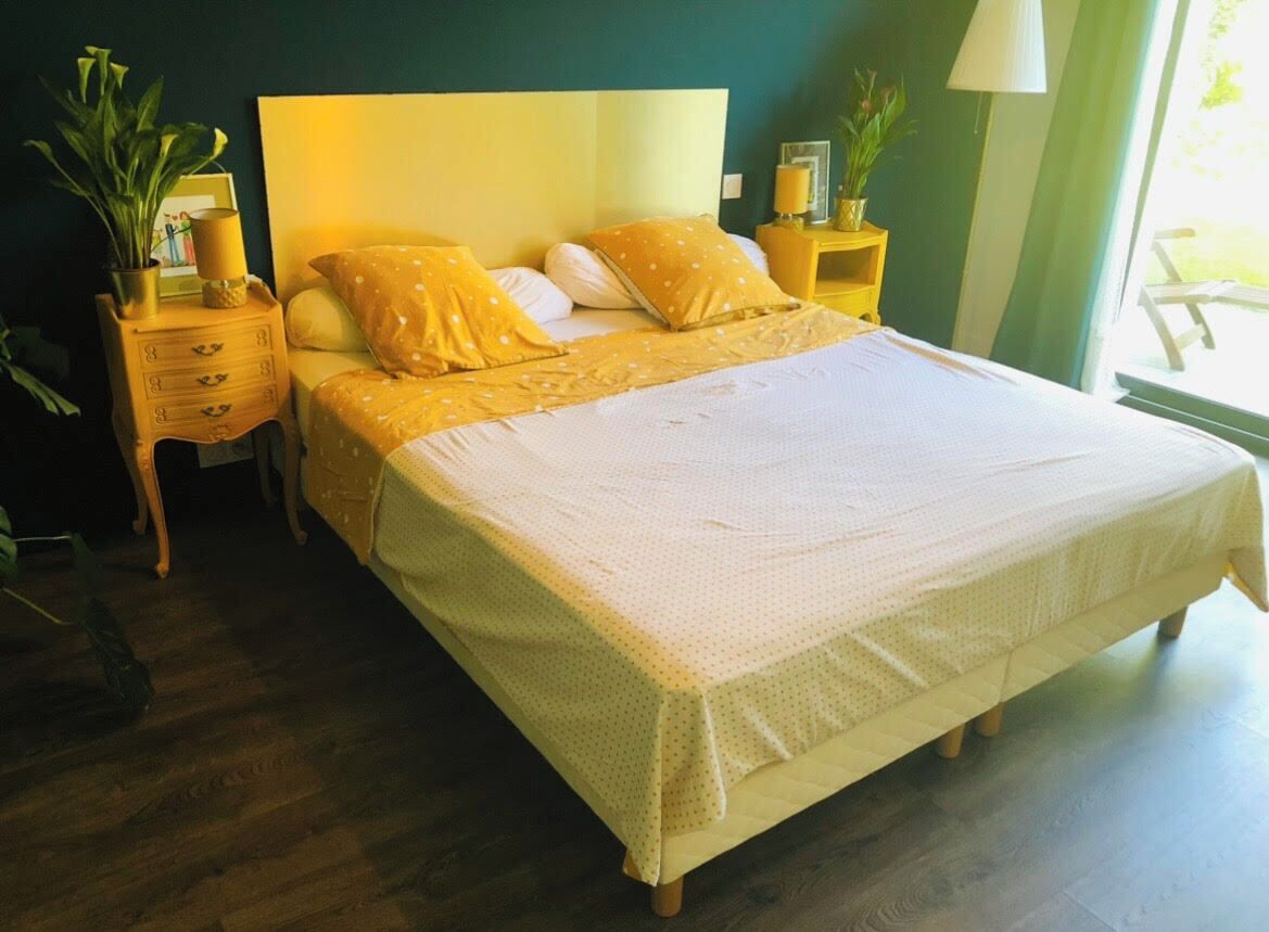 A bed with a white bedspread and yellow pillows, a wooden nightstand with a lamp, and a potted plant.