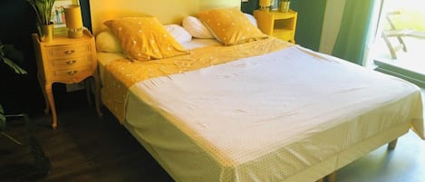 A bed with a white bedspread and yellow pillows, a wooden nightstand with a lamp, and a potted plant.