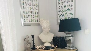 Ett hörnrum med ett mörkgrönt skåp, en byst, en inramad botanisk illustration, en lampa och en liten staty.