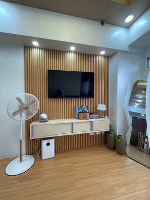 Un salon moderne avec un téléviseur à écran plat fixé à un mur en bois, un ventilateur sur pied, une petite poubelle et une console minimaliste agrémentée d’objets d écoratifs.