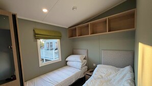 A single bed with a headboard, a bedside table, a window with a view of a house, and a shelf above the bed. - Haggerston Castle - Lakeside Retreat (Berwick-upon-Tweed)