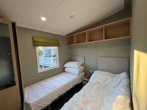 A single bed with a headboard, a bedside table, a window with a view of a house, and a shelf above the bed. - Haggerston Castle - Lakeside Retreat (Berwick-upon-Tweed)