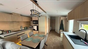 A compact kitchen with wooden cabinets, a dining area with a table and chairs, and a small window. - Haggerston Castle - Lakeside Retreat (Berwick-upon-Tweed)