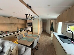 A compact kitchen with wooden cabinets, a dining area with a table and chairs, and a small window. - Haggerston Castle - Lakeside Retreat (Berwick-upon-Tweed)