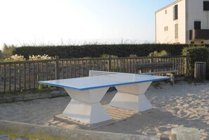 Ein Tischtennis-Tisch auf sandigem Untergrund, mit Holzzaun und einem Gebäude im Hintergrund.