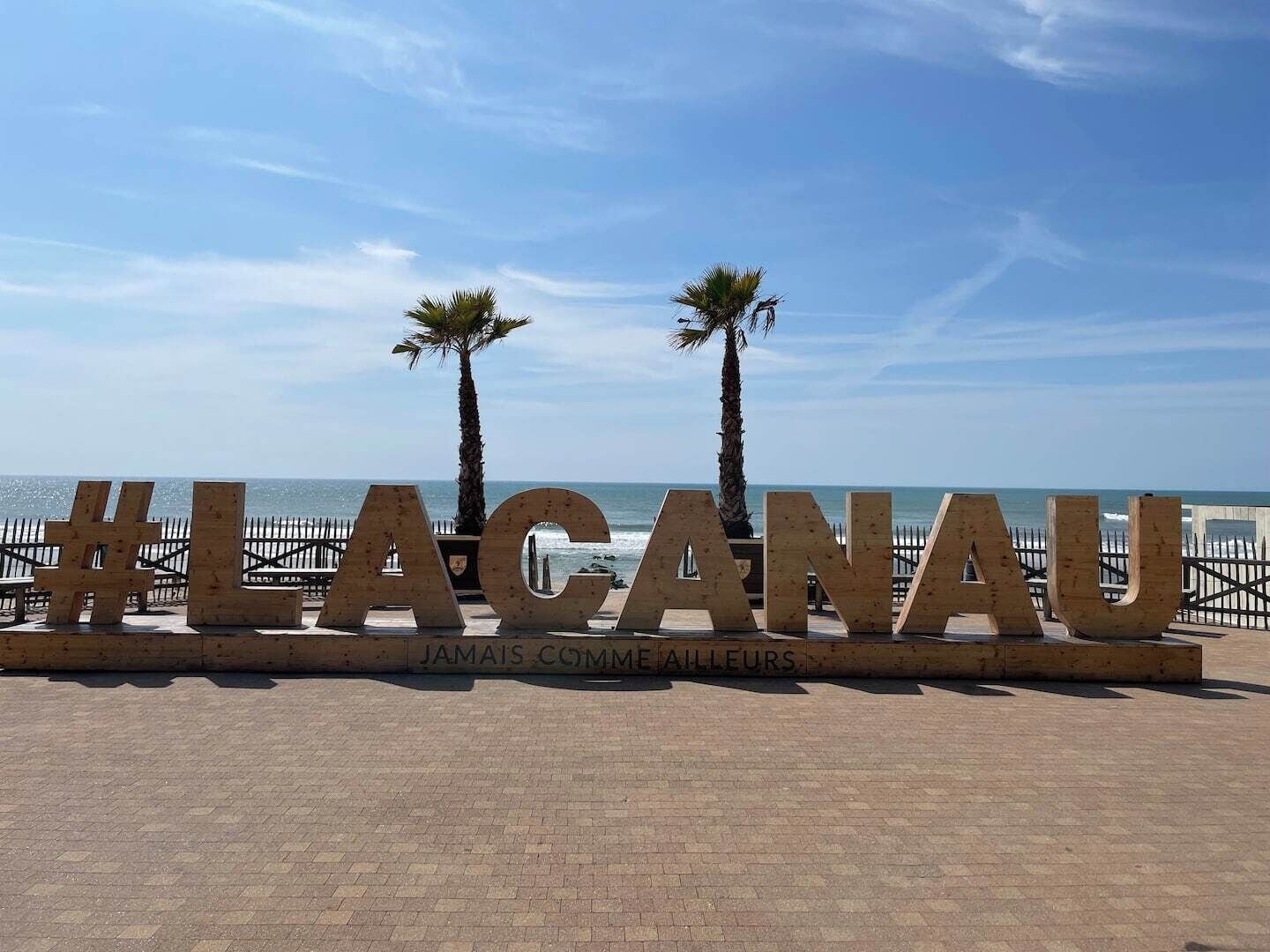 A wooden sign with the text '#LACANAU' and 'JAMAIS COMMUNE AILLEURS' against a backdrop of a beach and palm trees.