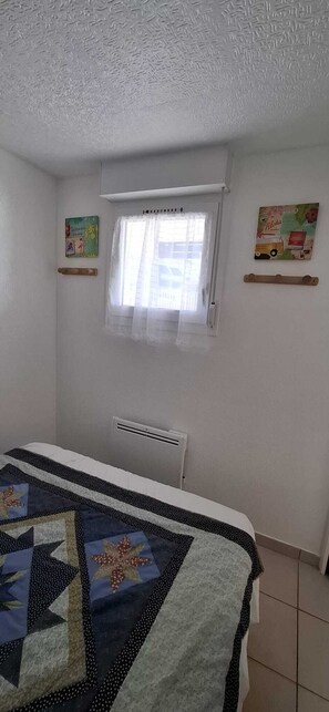 A bedroom with a bed, a window with a curtain, a radiator, and two wall decorations. - T2 RDC avec jardin - LACANAU OCÉAN (Lacanau)