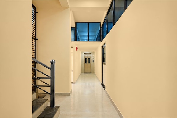 A corridor with a staircase on the left, a fire alarm on the wall, and an elevator at the end.