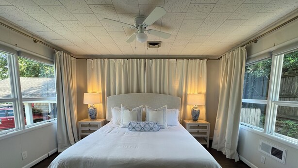 A bedroom with a large bed, two bedside tables with lamps, a ceiling fan, and windows with curtains. - Capstone Cottage | Walk to Bryant-Denny Stadium | 3 King Beds (Tuscaloosa)