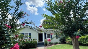 A house with a brick pathway leading to the entrance, surrounded by lush greenery and flowering plants. - Capstone Cottage | Walk to Bryant-Denny Stadium | 3 King Beds (Tuscaloosa)