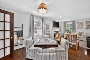 A dining area with a wooden table and striped chairs, a television above the fireplace, and a view of the outdoors through a window. - Capstone Cottage | Walk to Bryant-Denny Stadium | 3 King Beds (Tuscaloosa)