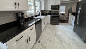 A modern kitchen with white cabinetry, a black countertop, stainless steel appliances, and a dining area with a sofa. - Capstone Cottage | Walk to Bryant-Denny Stadium | 3 King Beds (Tuscaloosa)