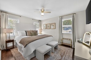 A bedroom with a bed, bedside table, bench, ceiling fan, window with curtains, and wall-mounted artwork. - Capstone Cottage | Walk to Bryant-Denny Stadium | 3 King Beds (Tuscaloosa)