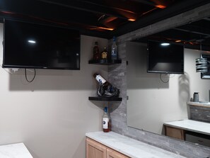 Un baño moderno con un espejo grande, dos televisores instalados en la pared y una encimera con una botella.