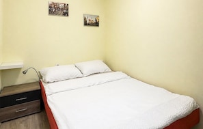Ein Einzelbett mit weißer Bettwäsche in einem Zimmer mit hellgelben Wänden, einem Nachttisch und zwei kleinen Bildern an der Wand.