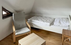 Un petit logement composé d’une seule pièce, avec un plafond en pente, une chaise en bois, une table en bois et un lit avec du linge de lit blanc.