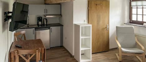 A compact room with a kitchenette, a wooden door, a white shelving unit, a cushioned chair, and a bed.
