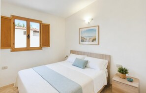 Ein Schlafzimmer mit einem Bett, einem Nachttisch, einer Wandleuchte, einem Fenster mit Holzläden und einem gerahmten Bild an der Wand.