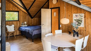 Ett rum med en säng, ett matbord med stolar, en vit stol med en mjuk kudde, ett träpanelat tak och ett fönster med utsikt över grönområden.