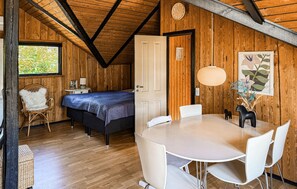 A room with a bed, a dining table with chairs, a white chair with a fluffy cushion, a wooden ceiling, and a window with a view of greenery. - Amazing home in Rødby with WiFi (Rødby)