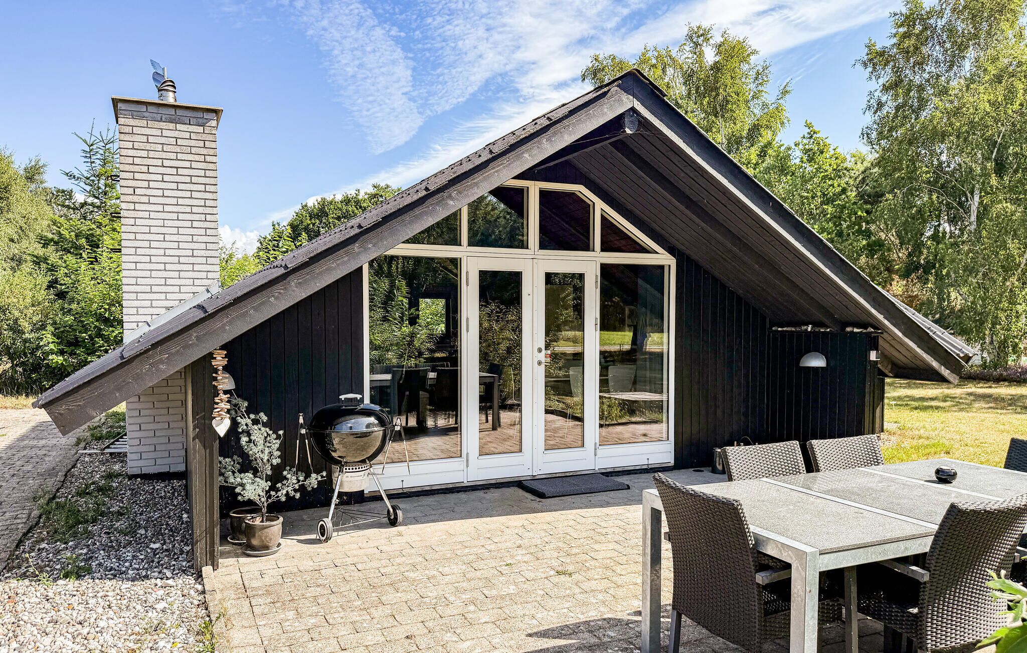 Une maison moderne avec un toit en pente, une cheminée en briques et une terrasse dotée d’une table et de chaises.