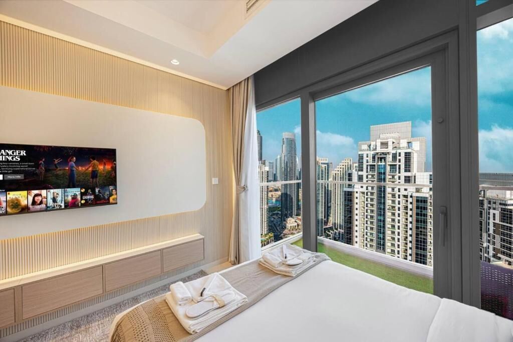 A modern hotel room with a large bed, a flat-screen TV mounted on the wall, and a city view through large windows.