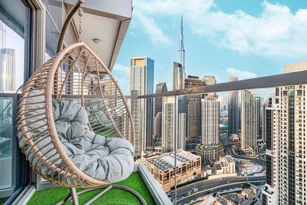 Interior de um arranha-céu com uma cadeira suspensa, vista para a cidade e piso de grama artificial verde.
