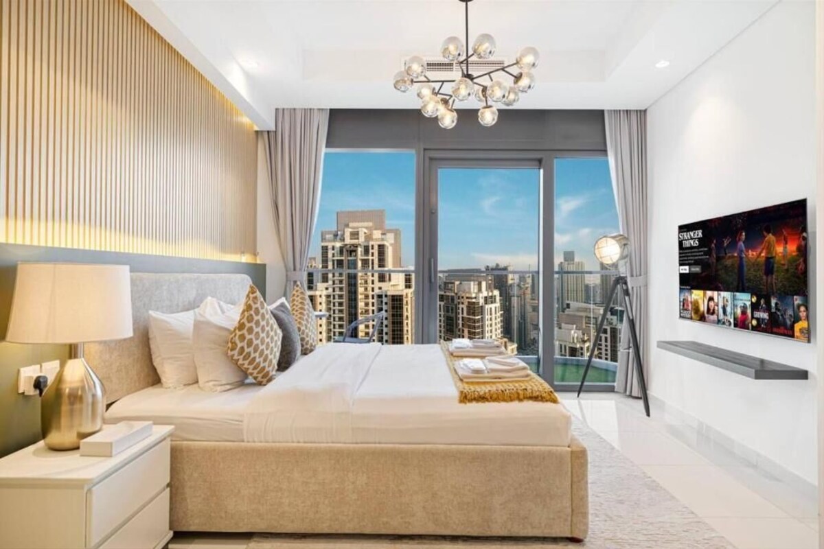Ein modernes Hotelzimmer mit einem großen Bett, Blick auf die Stadt, einer Kandelaberleuchte und einem an der Wand befestigten Fernseher.