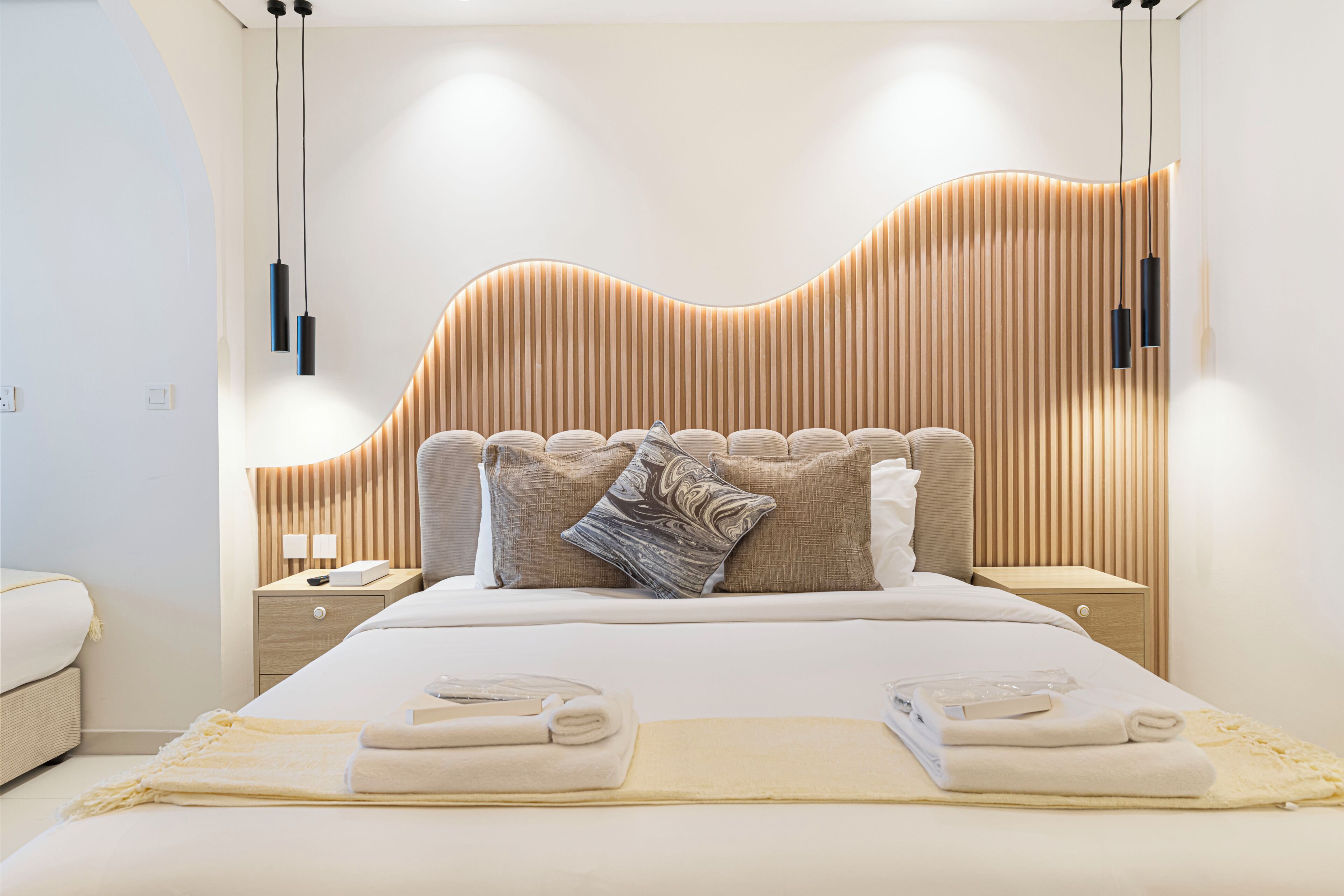 Ein modernes Schlafzimmer mit einem gebogenen Kopfteil, einem ordentlich bezogenen Bett mit Handtüchern und Nachttischlampen.