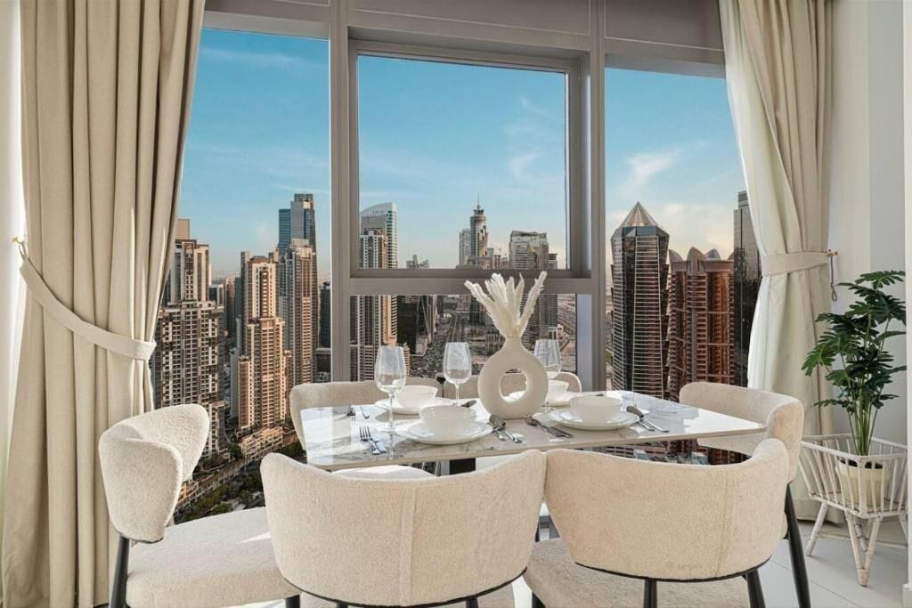 Uma sala de jantar com mesa posta para quatro pessoas, amplas janelas com vista para a cidade e um vaso de plantas ao lado.