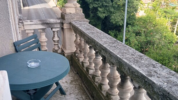 A balcony with a table and chairs, a stone balustrade, and a view of a street and trees. - Hostel veli jože (Pula)