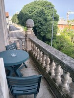 Un balcón con mesa y sillas, baranda de piedra y vista a la calle y árboles.