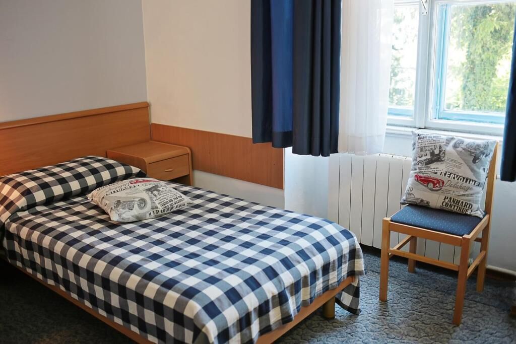 A single bed with a checkered bedspread, a nightstand, a chair with a cushion, a window with curtains, and a radiator.