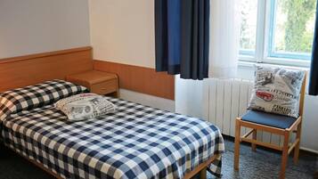 A single bed with a checkered bedspread, a nightstand, a chair with a cushion, a window with curtains, and a radiator.