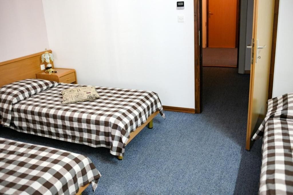 A hotel room with a checkered bedspread, a bedside table with a lamp, and a door leading to another room.