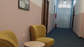 Ein Flur mit zwei gelben Sesseln und einem kleinen runden Tisch, einem gerahmten Infoposter an der Wand und einer Tür am Ende.