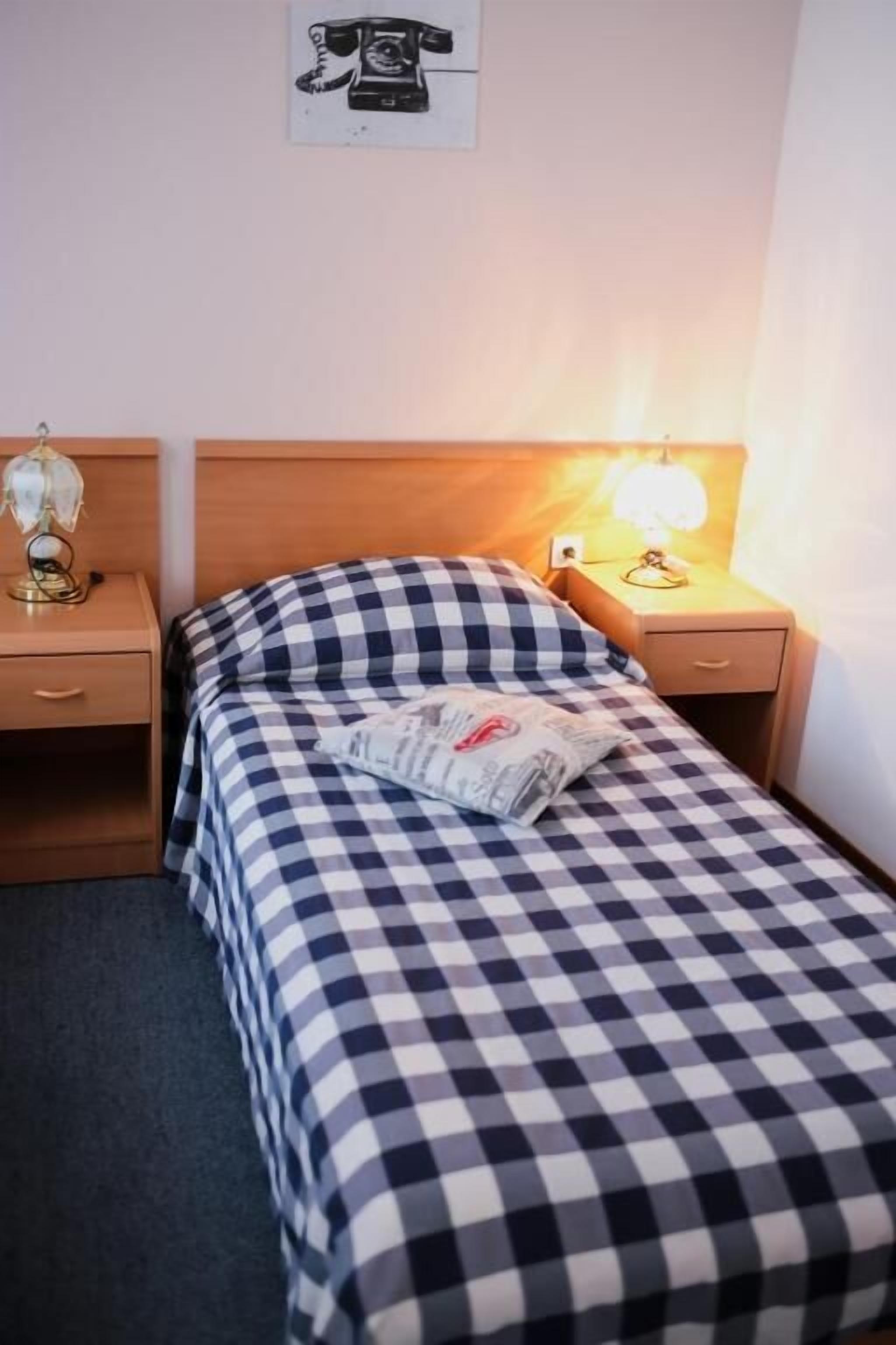 A single bed with a checkered bedspread, two bedside tables with lamps, a wall-mounted telephone, and a small decorative item on the left table.