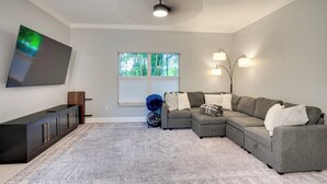A living room with a sectional sofa, a flat-screen TV mounted on the wall, a window with blinds, and a ceiling fan. - Heated Pool & Lanai Cookouts! Cape Coral Getaway (Cape Coral)