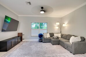 A living room with a sectional sofa, a flat-screen TV mounted on the wall, a window with blinds, and a ceiling fan. - Heated Pool & Lanai Cookouts! Cape Coral Getaway (Cape Coral)