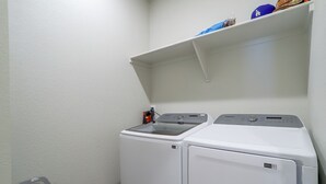 A laundry room with two white washers and dryers, a small shelf above the dryers, and a gray laundry basket against the wall. - Private Yard w/ Patio! Modern Commerce City Gem (Commerce City)