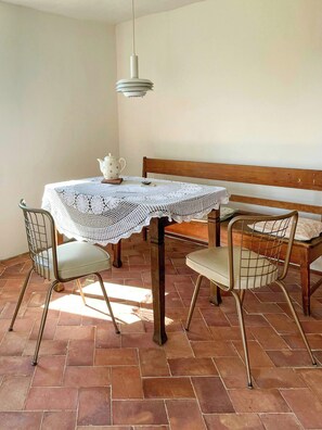 A dining area with a table covered by a lace tablecloth, two wireframe chairs, a bench, and a hanging light fixture. - Quiet studio retreat with a peaceful garden (Wenzlow OT Boecke)