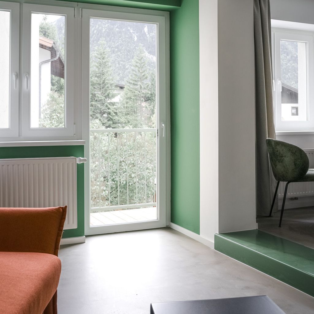 Une pièce avec un mur vert, un radiateur blanc, une porte-fenêtre coulissante donnant sur un balcon et une chaise verte.