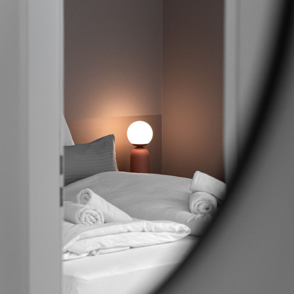 A neatly made bed with white linens and a round table lamp in the background.