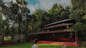 A person walking on a stone pathway towards a building with a red roof, surrounded by lush greenery and trees. - Native Retreats Varkala (Hariharapuram)