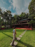 Una persona camina por un sendero de piedra hacia un edificio de techo rojo, rodeado de abundante vegetación y árboles.