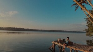 A wooden dock extending into calm water with a person sitting on it, palm trees in the background, and a clear sky. - Native Retreats Varkala (Hariharapuram)