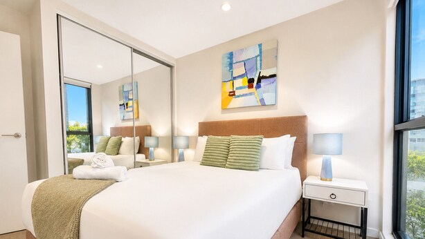 Quarto de hotel com uma cama grande, uma mesinha de cabeceira com abajur, um espelho e uma janela com vista.